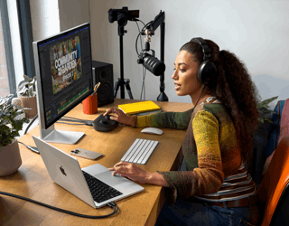 Una mujer en su casa trabajando desde su escritorio con una MacBook Pro conectada a dos monitores externos, usando audífonos y hablando en un micrófono
