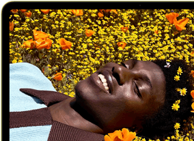 MacBook Neo, color amarillo cítrico, la pantalla muestra a una persona sonriente acostada en un campo de flores bajo el sol